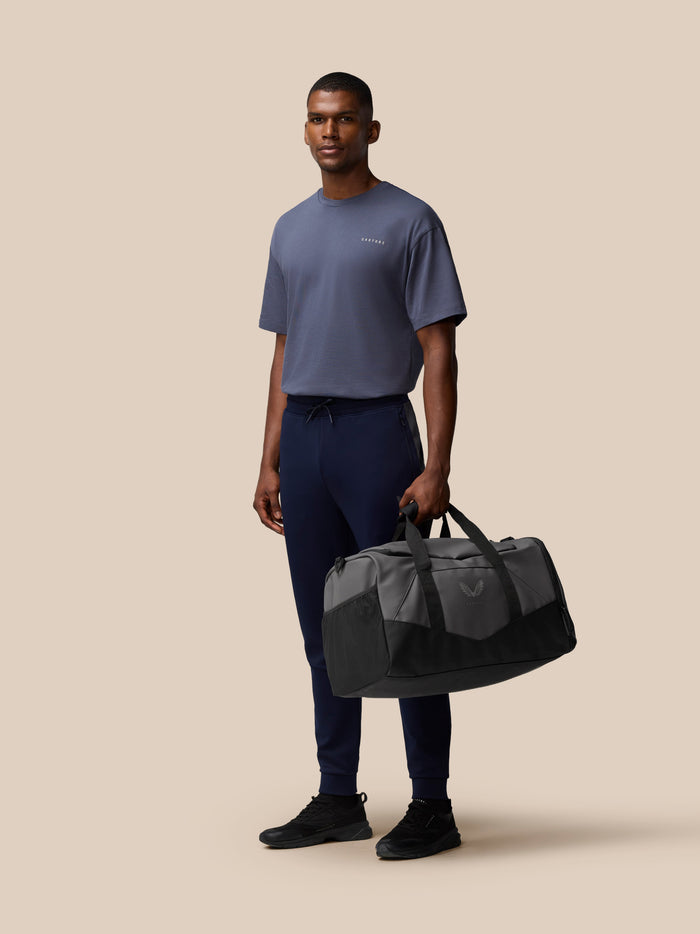 Person standing with a grey Core Holdall in the foreground, wearing a blue gray T shirt and navy pants