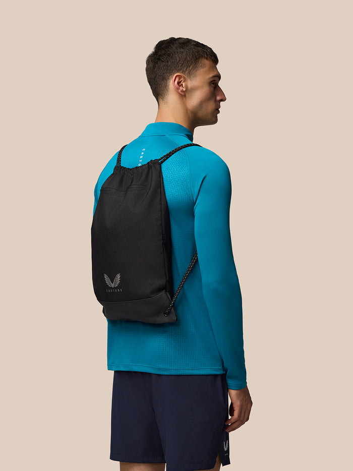 Side view of a man wearing a black Castore drawstring gym bag as a backpack over a bright teal athletic shirt