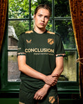 Portrait of a woman modeling the FC Utrecht 25/26 women's away shirt in dark green with tan shoulder panels and crest.