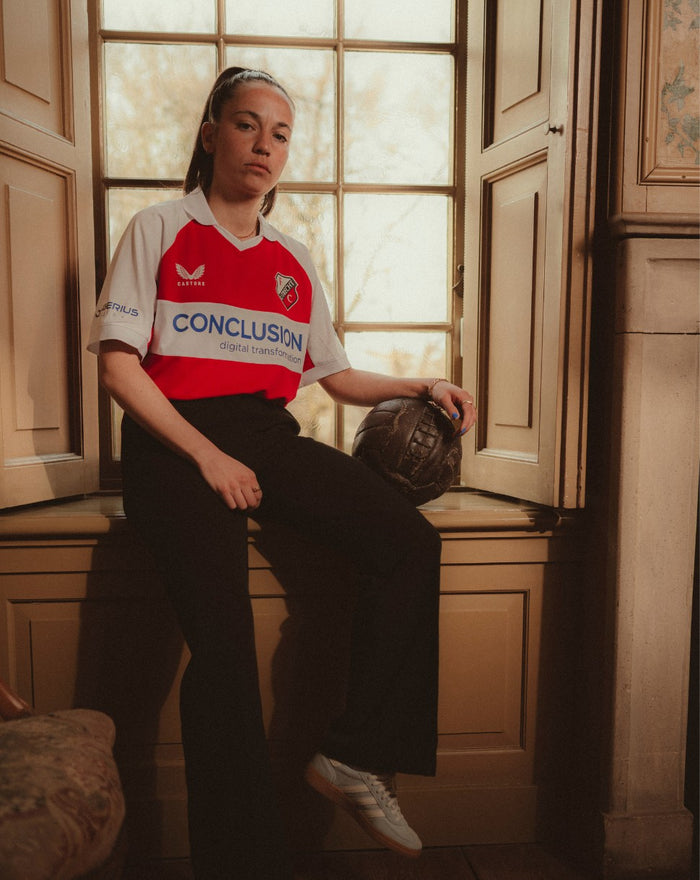 Vrouwelijk model draagt FC Utrecht Vierde Shirt 24/25 met V-hals en clublogo links op de borst in een kamer met raam en voetbal